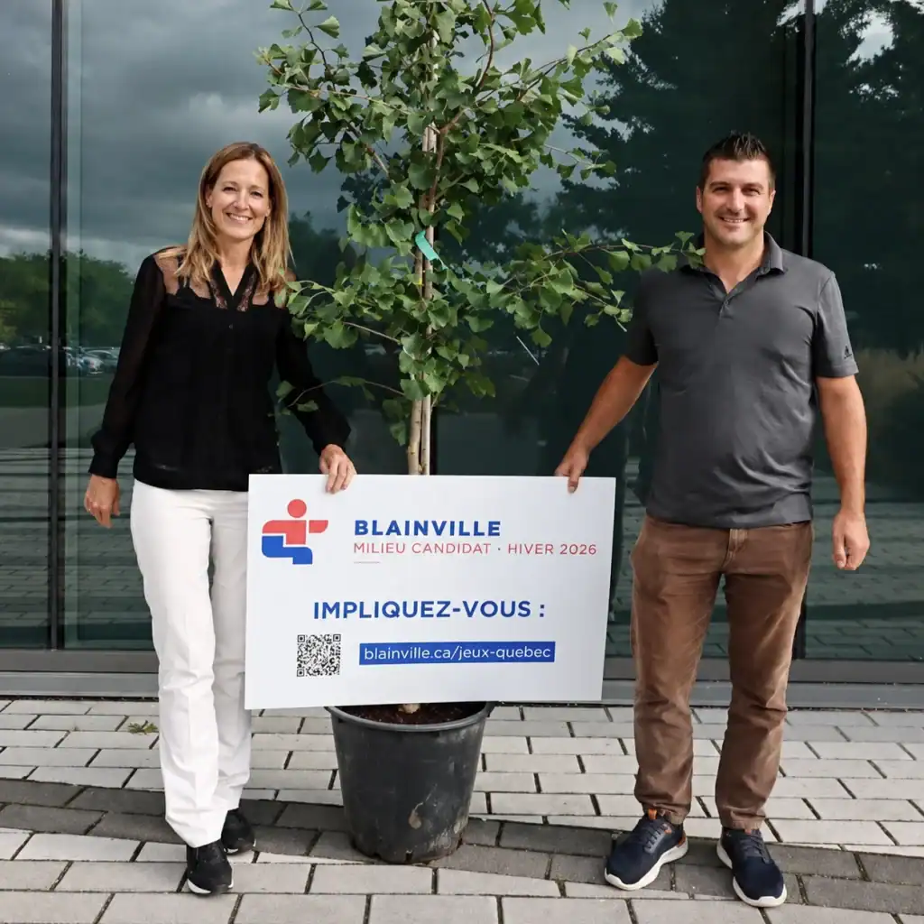 Deux personnes posent avec un arbre en pot devant un bâtiment moderne.