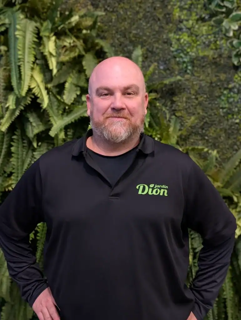 Homme en polo noir avec logo Jardin Dion devant un mur de plantes vertes.