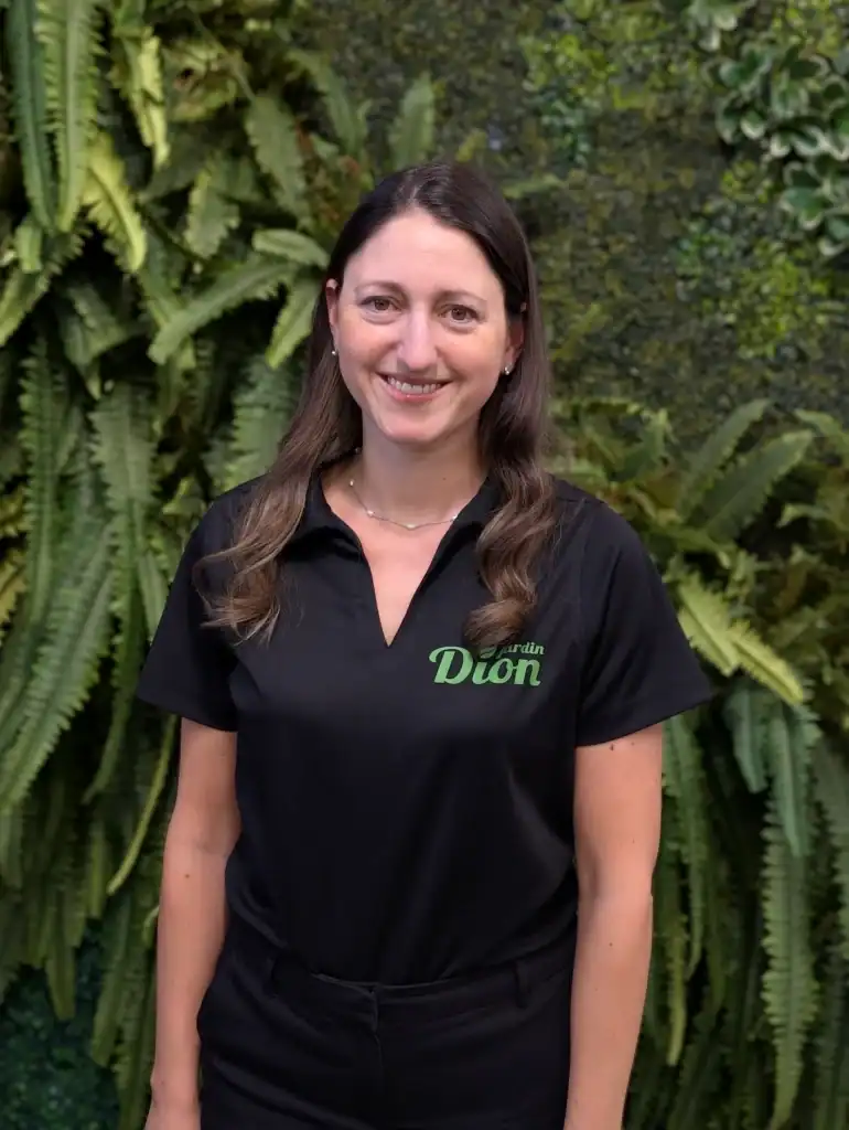 Femme souriante en uniforme Jardin Dion devant un mur végétal luxuriant.